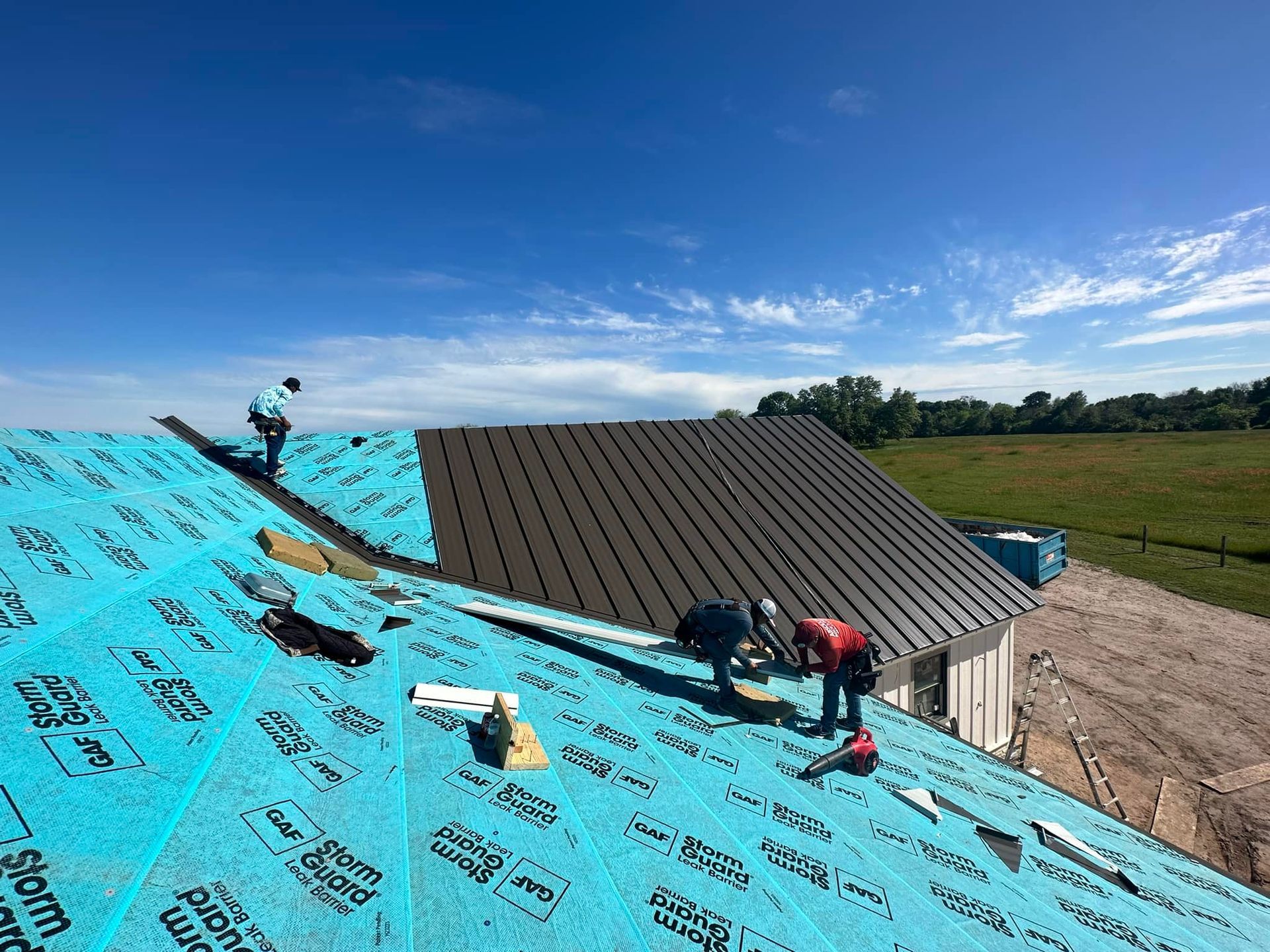 Roofers installing dark metal roofing on a house under a blue sky.
