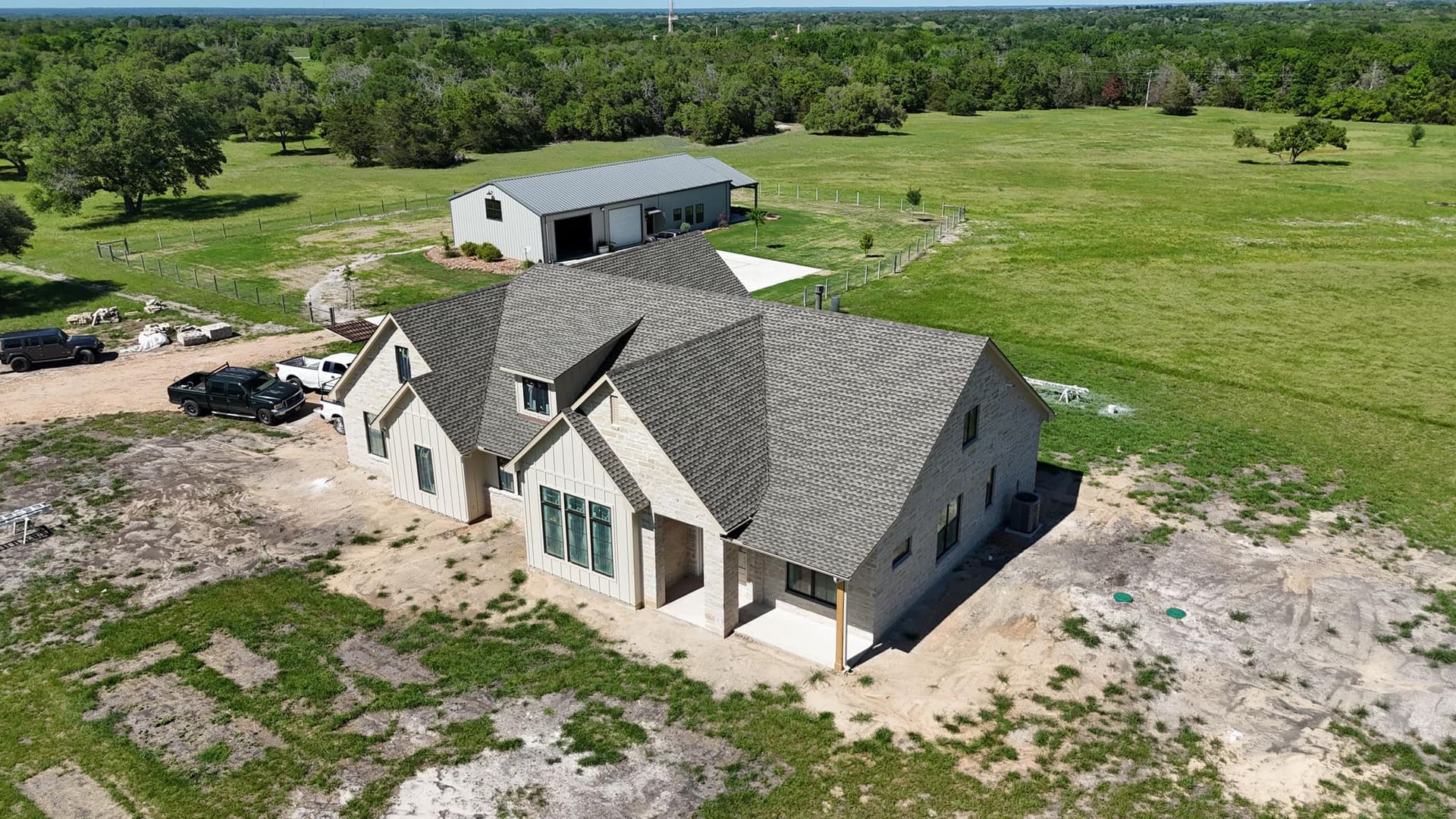Newly constructed house with gray roof, set on grassy land, with a barn in the background.