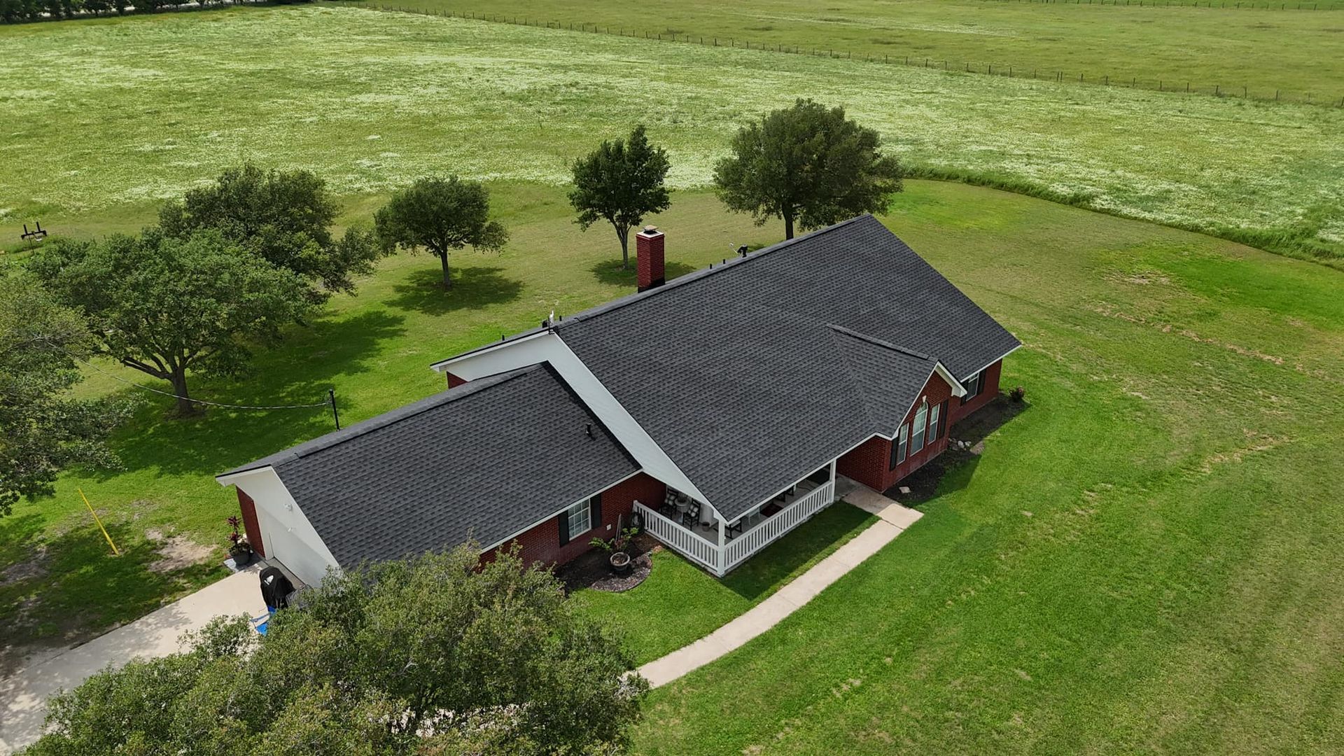 Aerial view of a red house with a dark roof on a green lawn, surrounded by trees and a field of white flowers.