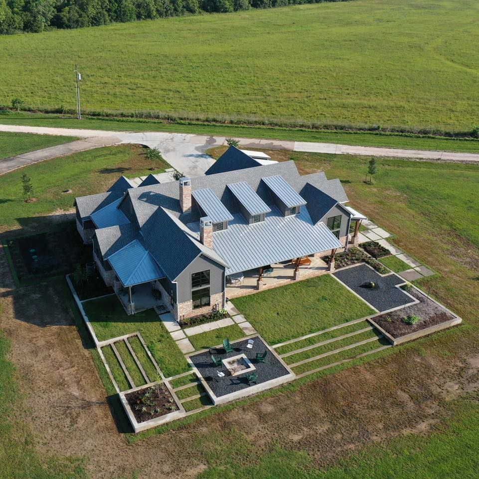 Aerial view of a modern farmhouse with a blue roof, surrounded by green grass and a gravel driveway.