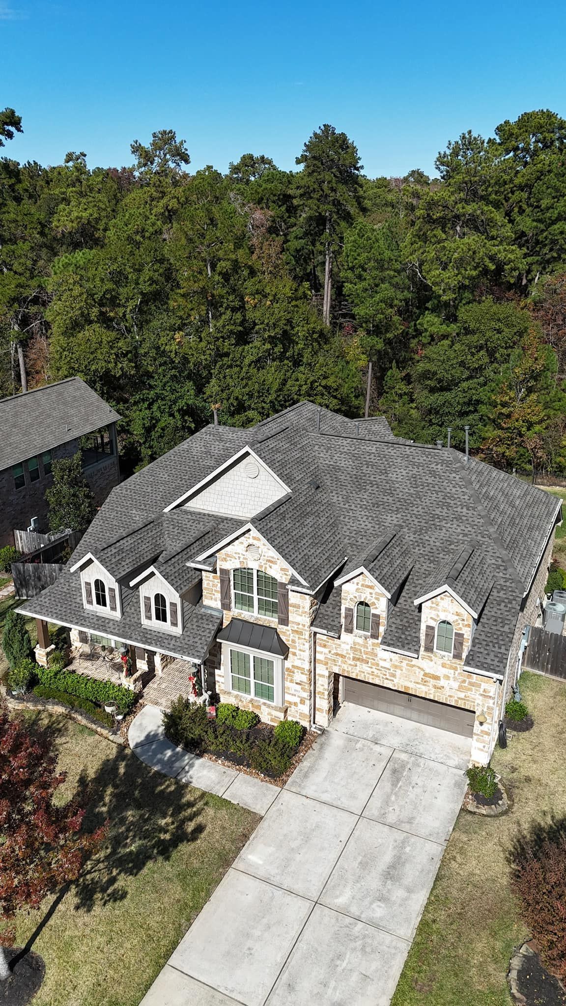 House with a stone facade, gray roof, and long driveway, surrounded by trees.