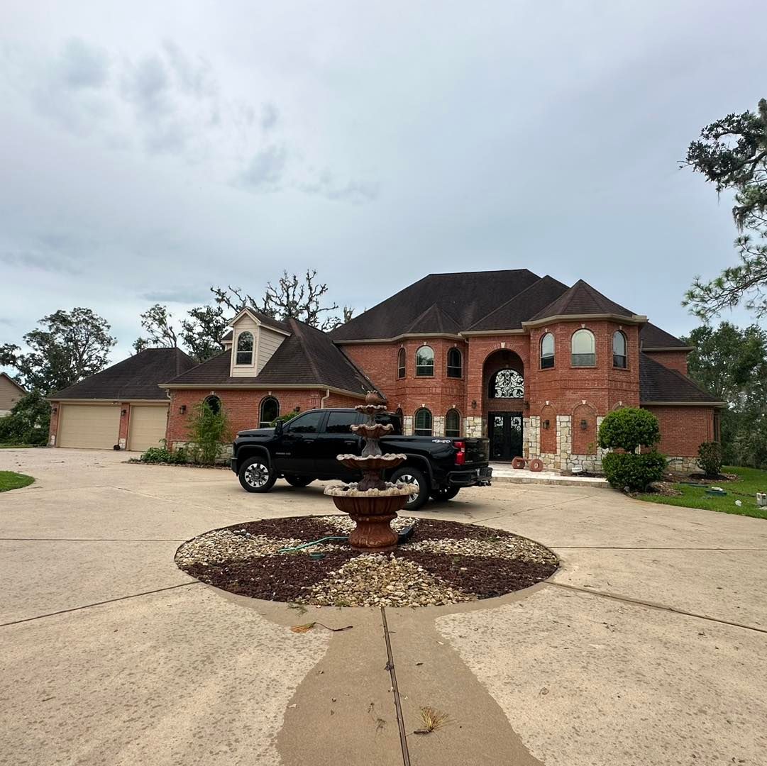 Large brick house with black truck parked on circular driveway. Fountain in center. Overcast sky.
