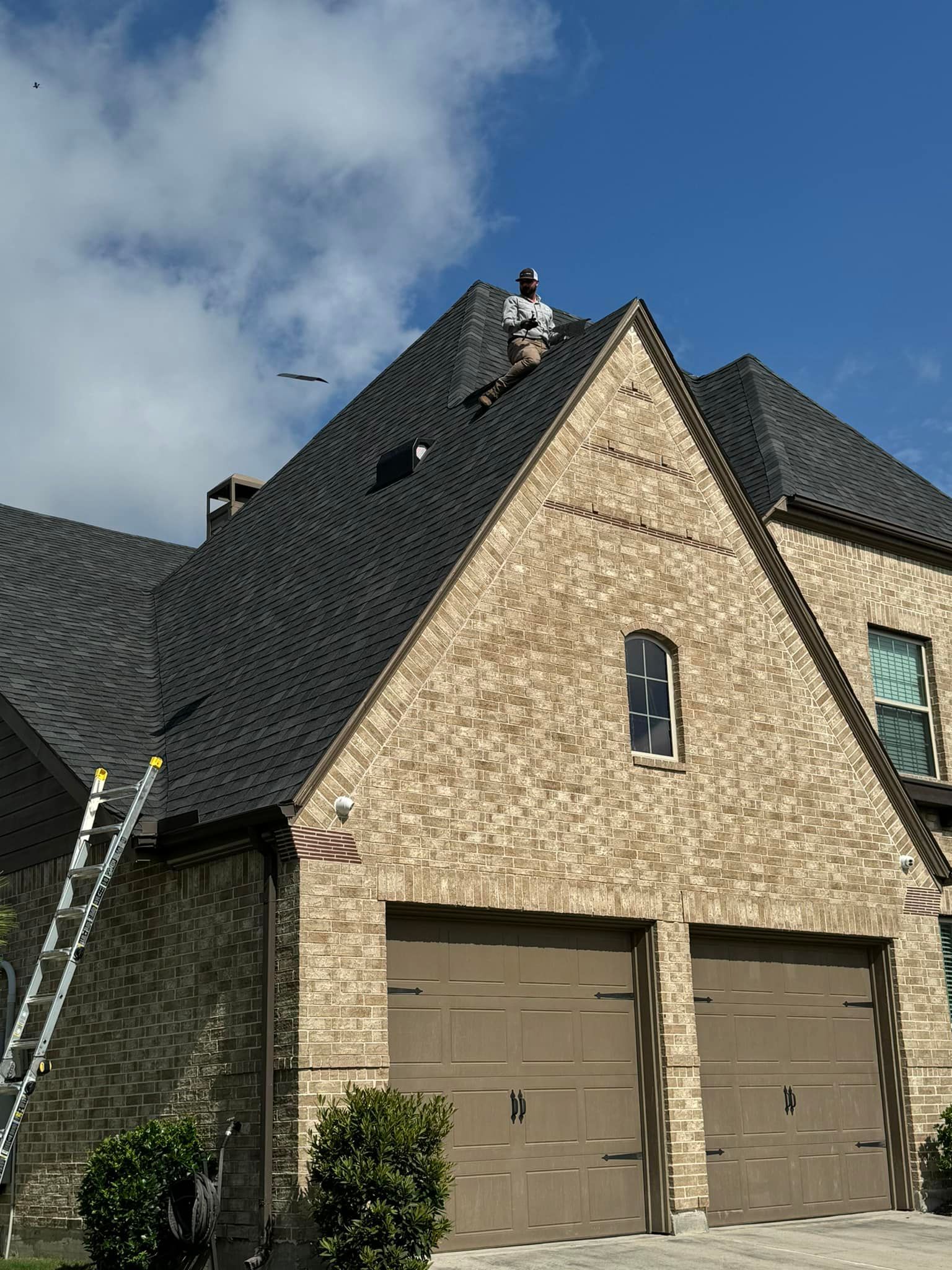 Person on roof installing shingles on a brick house with a garage door and ladder.