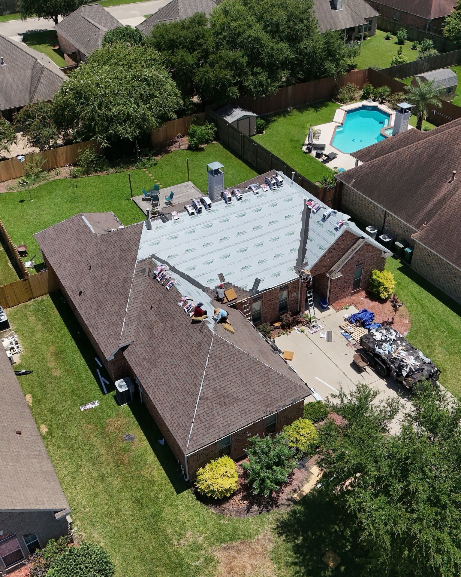 Aerial view of a house with roofing work in progress, workers on roof, blue tarp, tools, and a swimming pool.