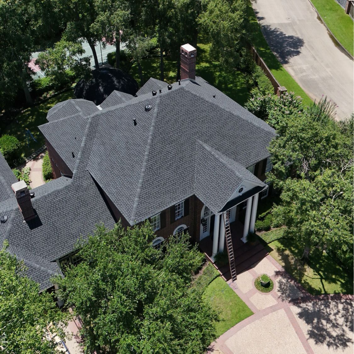Aerial view of a large house with a dark roof, surrounded by trees and a brick driveway.