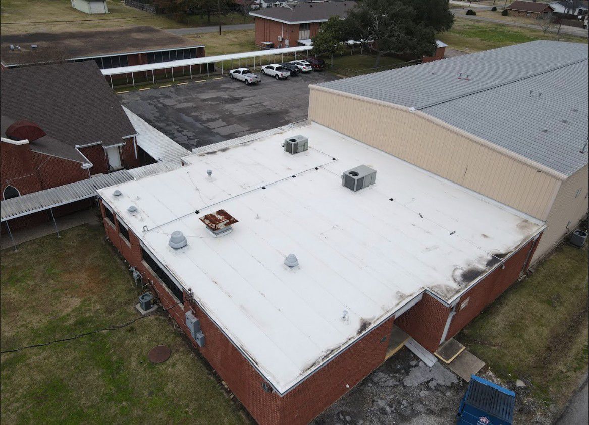 Aerial view of a white commercial roof with several air conditioning units; brick building with other buildings nearby; overcast sky.