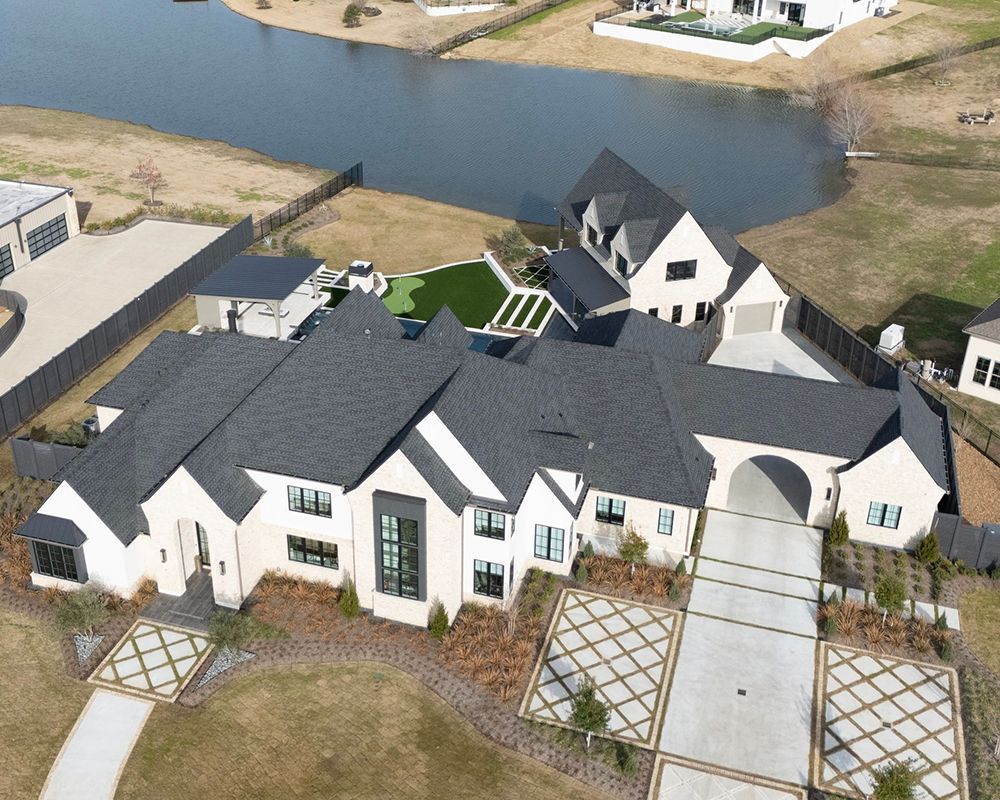 Aerial view of a large, modern white house with a dark roof and a lake in the background.