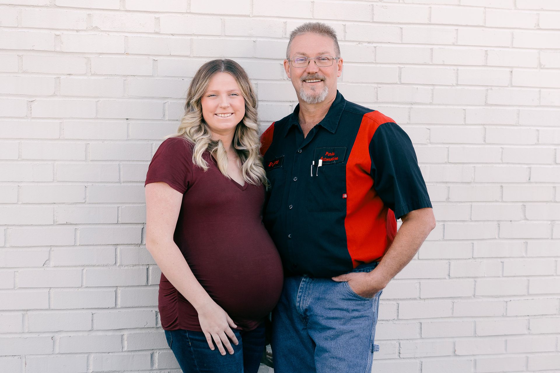 Pregnant woman and man standing in front of a white brick wall. The woman wears a burgundy shirt; the man wears a red and black shirt.