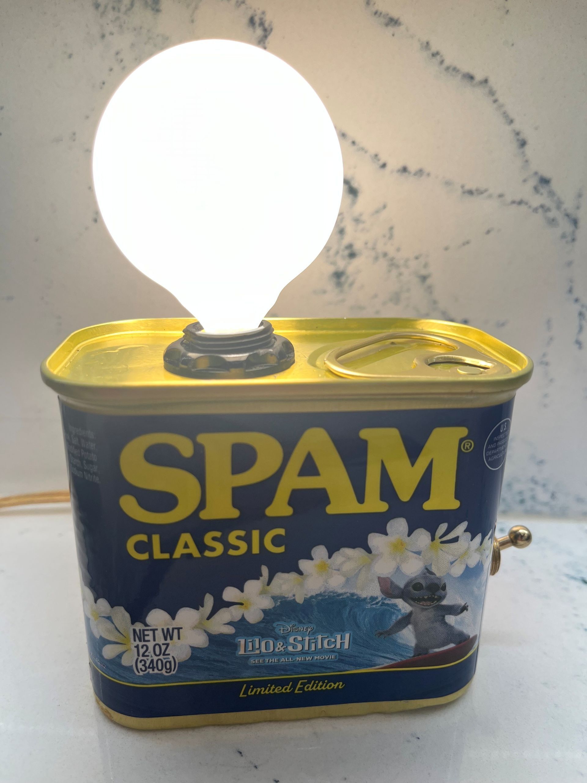 A Spam can lamp with a lit white bulb on top. The can is blue with yellow text and a Hawaiian print.