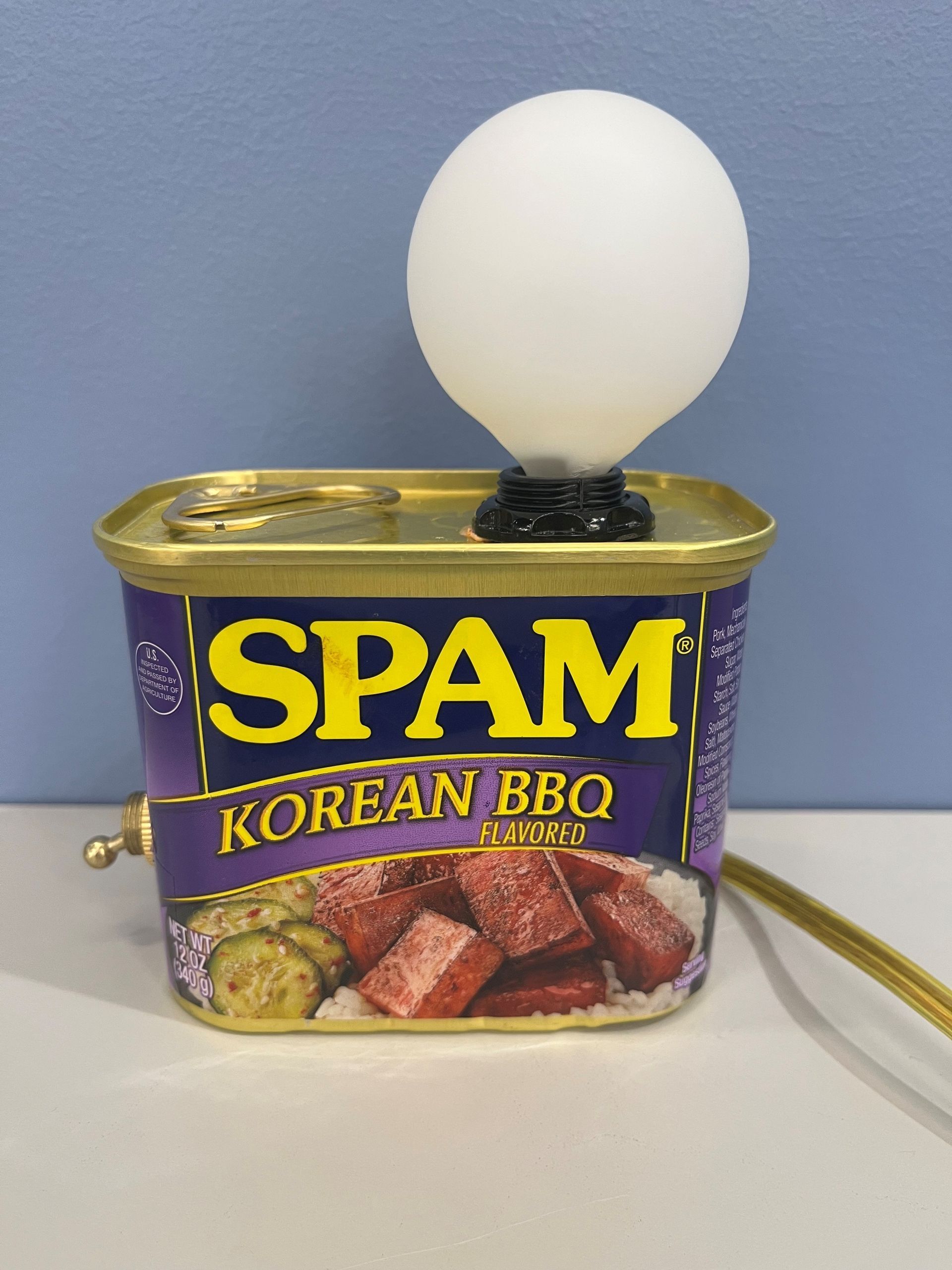 A Spam can repurposed as a lamp, with a lightbulb on top and a golden rod for a cord, on a white surface.