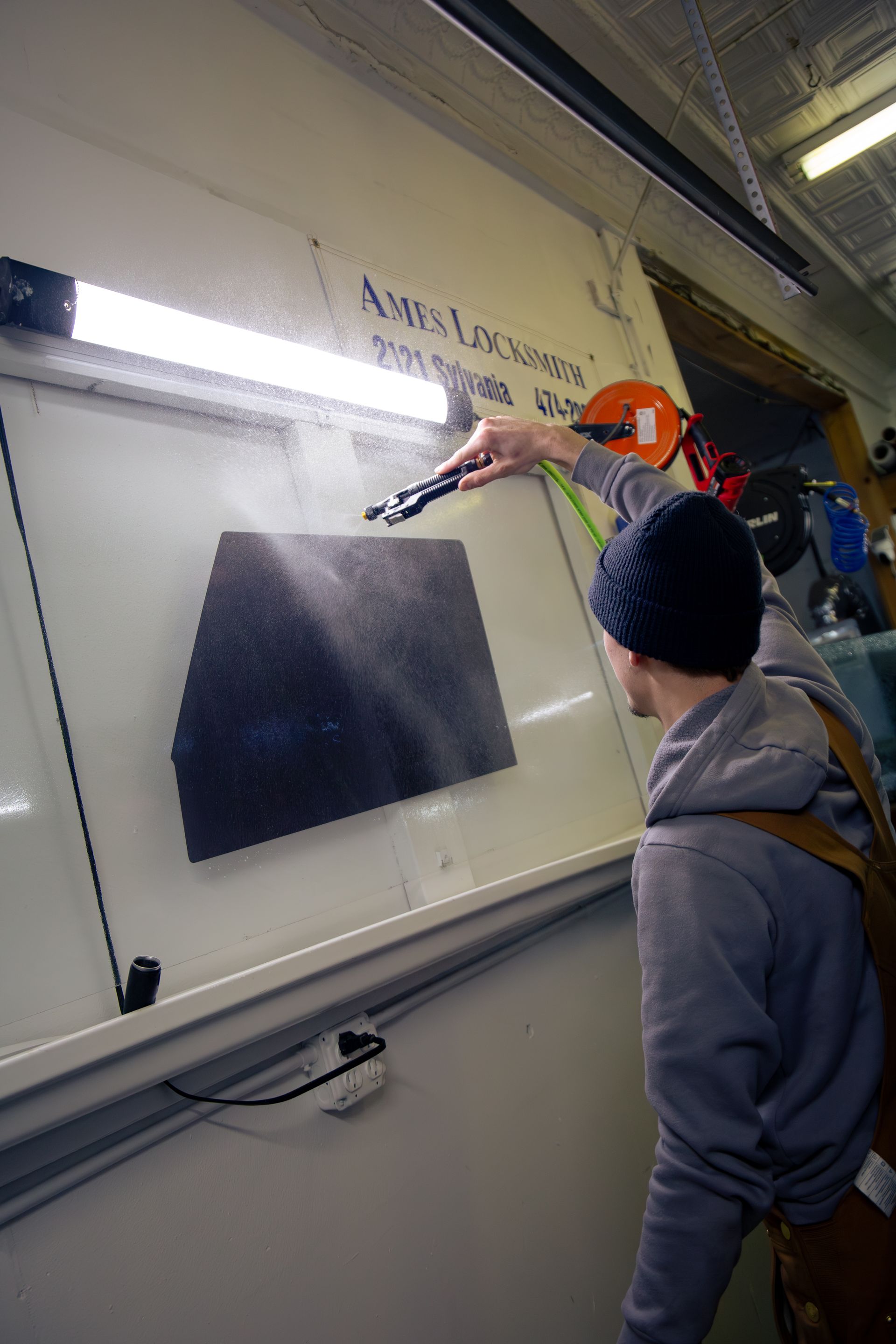 Person spraying a dark square object mounted on a white surface, possibly for a project or experiment, in a workshop.