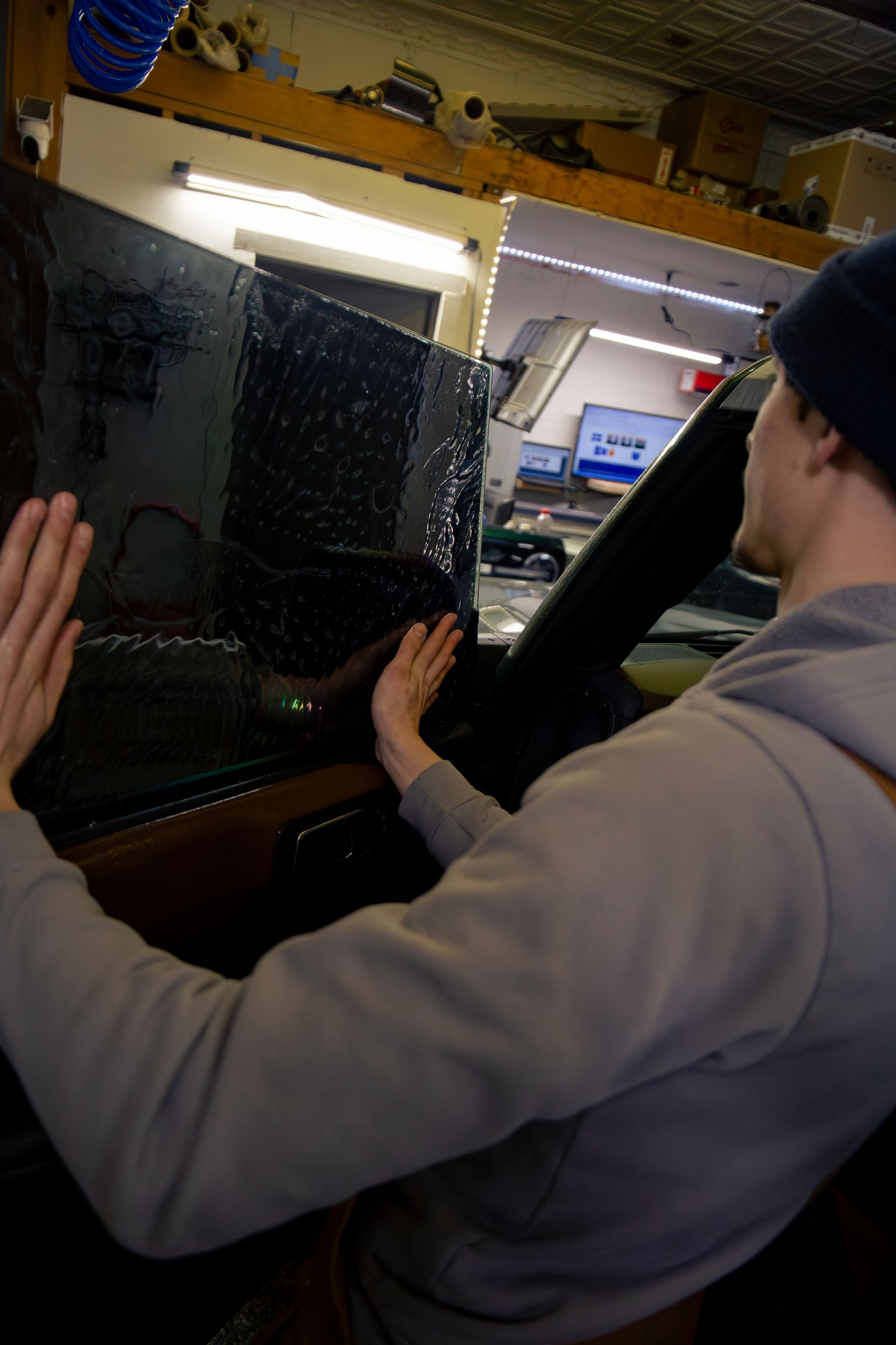 Person applying tint to car window in garage, hands flat against dark film.