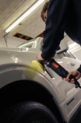A person uses a yellow buffing pad on a power tool to polish the side panel of a white vehicle in a workshop.