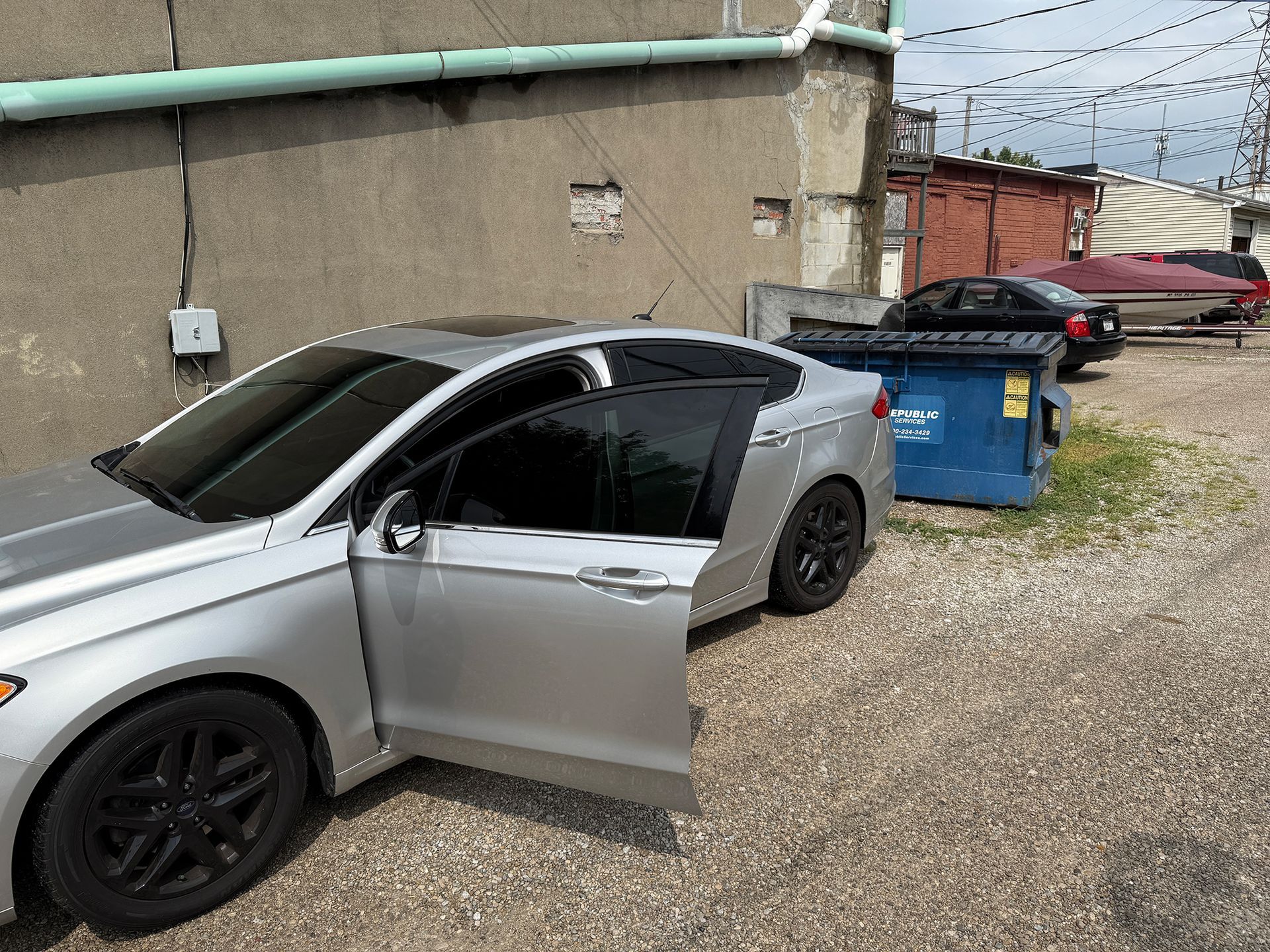 Silver car with open door parked next to a dumpster in an alley.