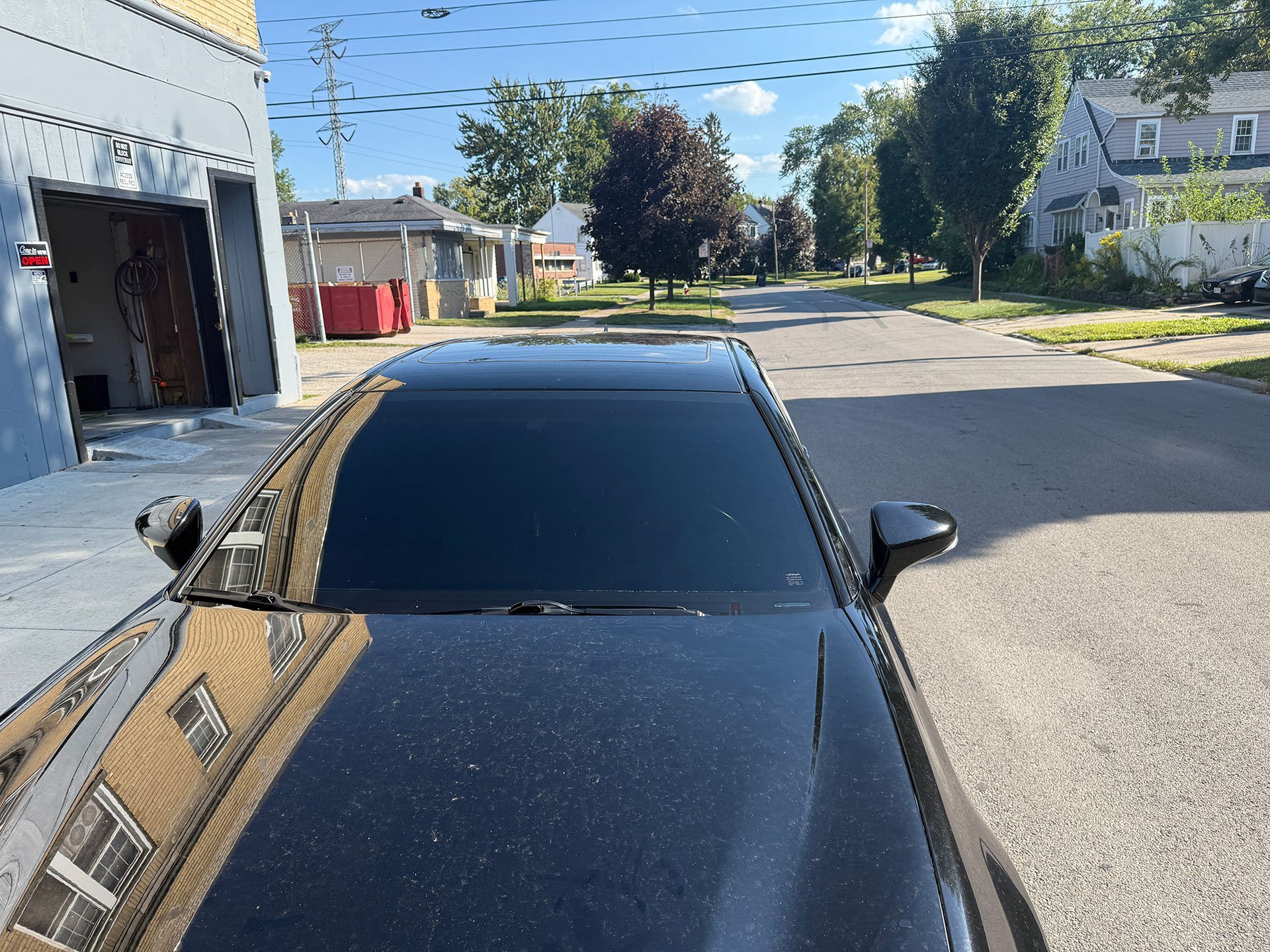 Black car parked on street, front end in view. Buildings and trees in background under a sunny sky.
