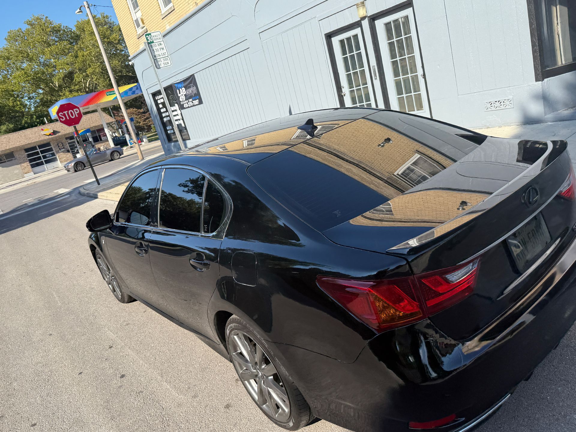 Black Lexus sedan parked on a city street. Building and stop sign visible in the background.