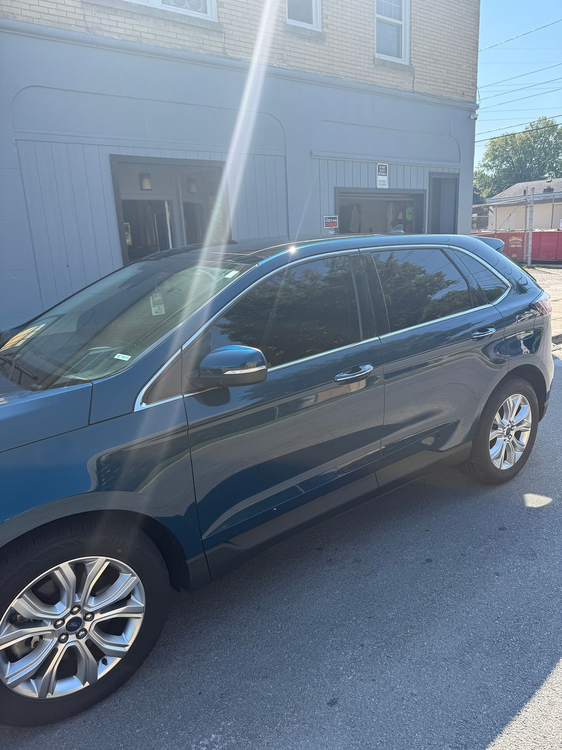 Dark teal SUV parked outside a light blue building, with heavily tinted windows. Sunlight present.