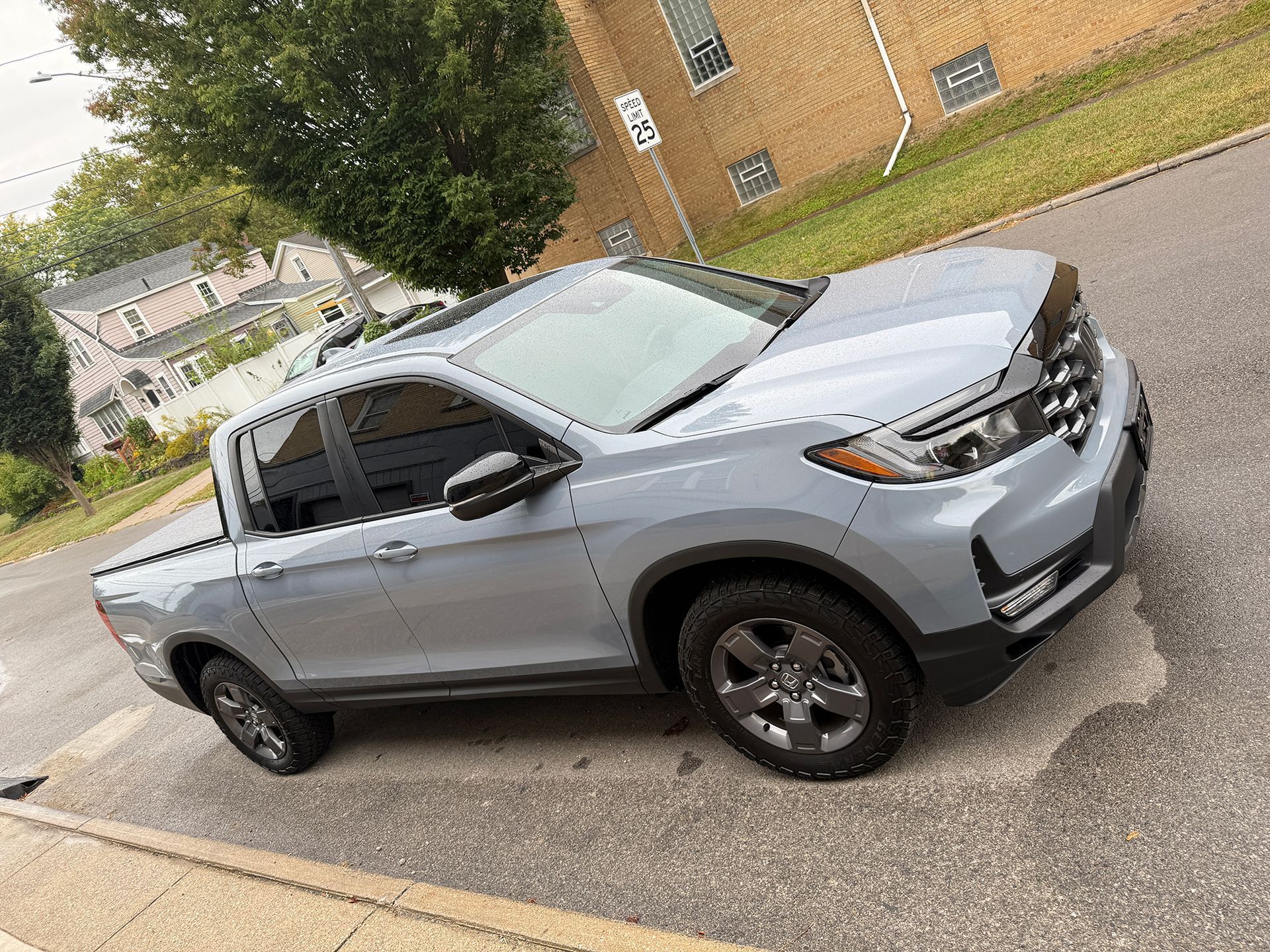 Gray Honda Ridgeline pickup truck parked on a street near a grassy area and brick building.