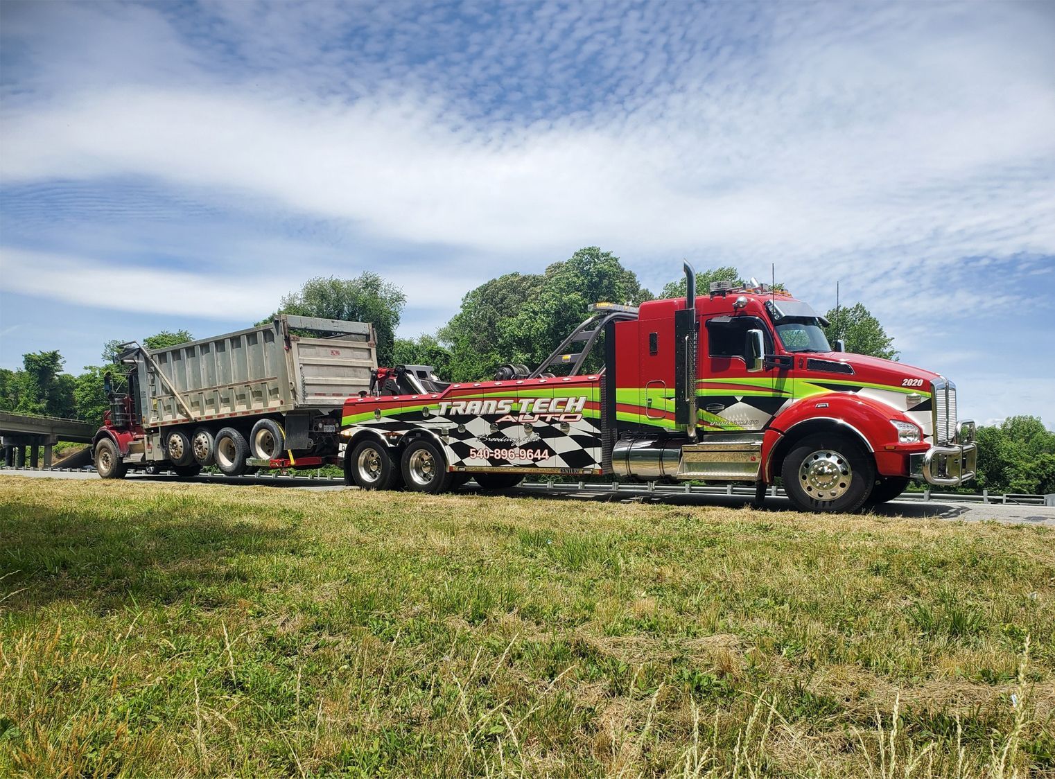 Red tow truck hauling a dump trailer along a grassy roadside under a cloudy sky