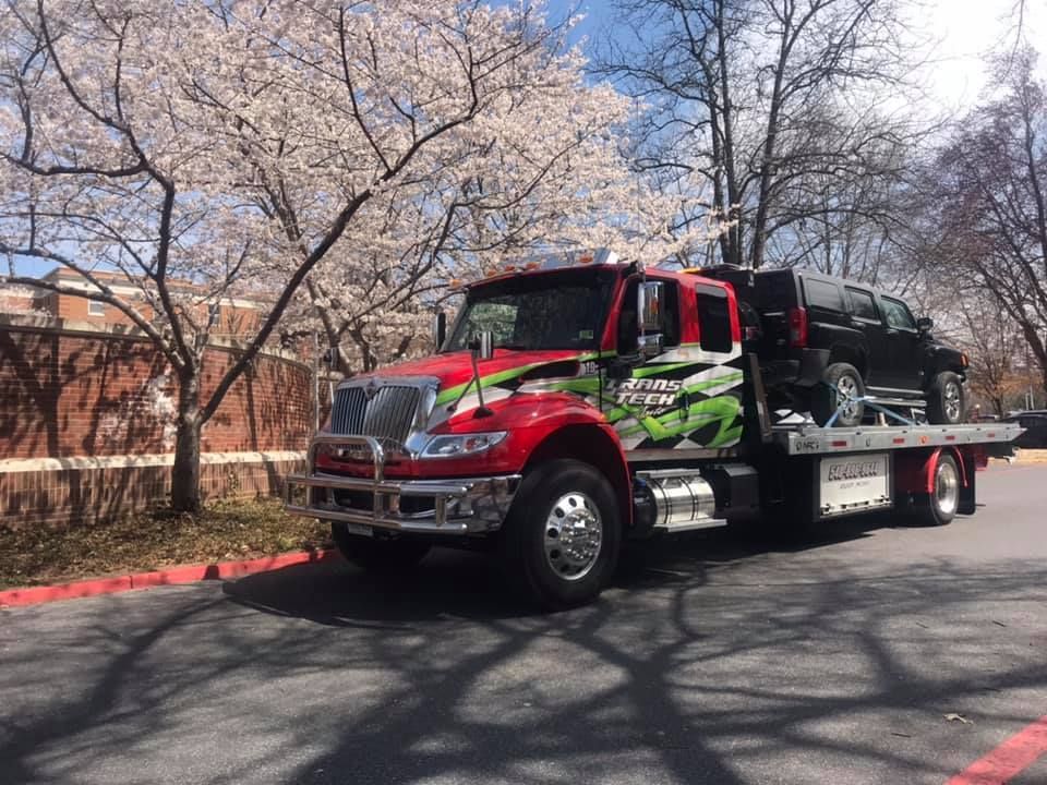 Red tow truck carrying a black car on a flatbed, parked beside blooming trees.