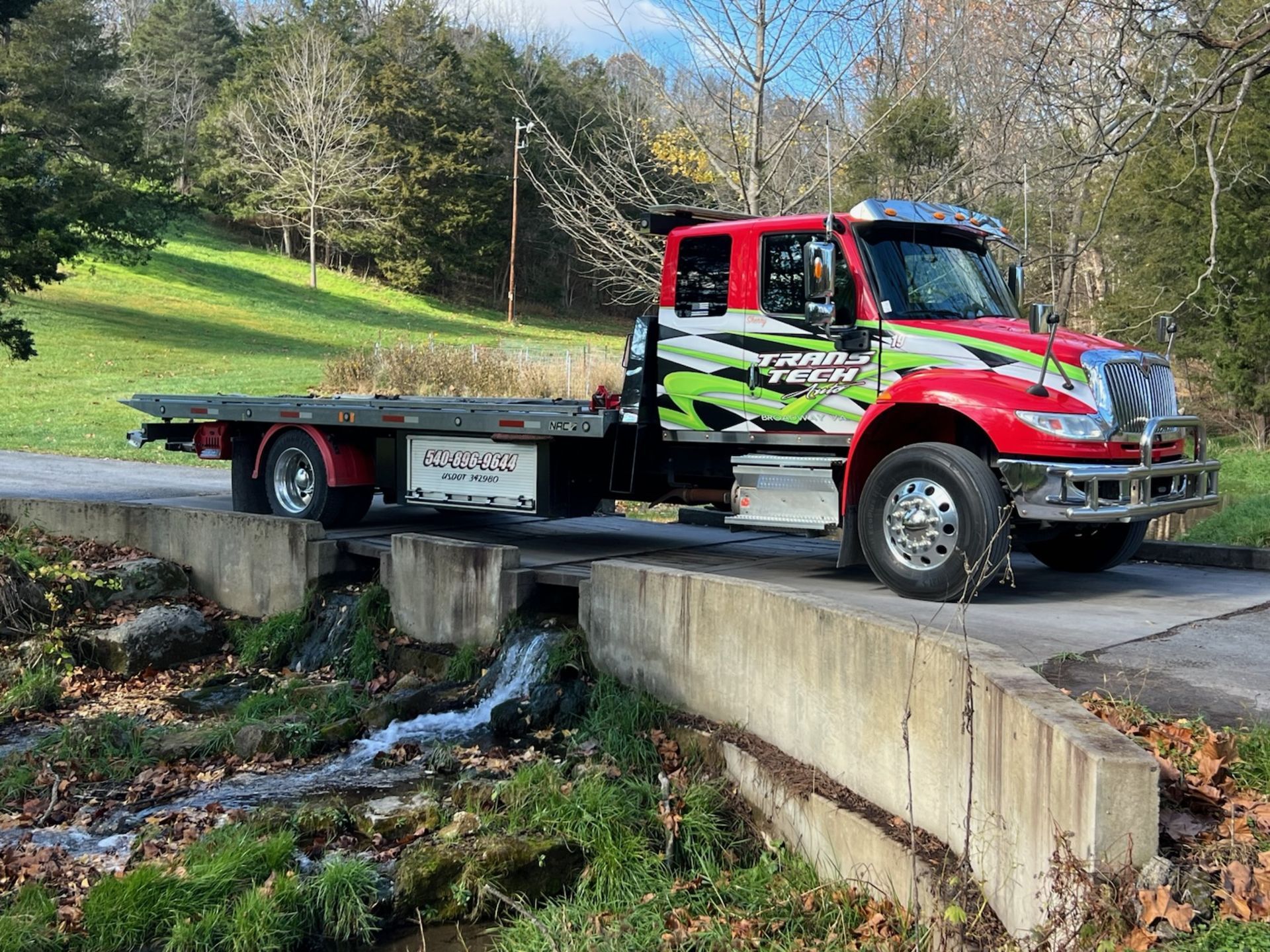 Red flatbed tow truck crossing a small concrete bridge over a creek in a wooded area