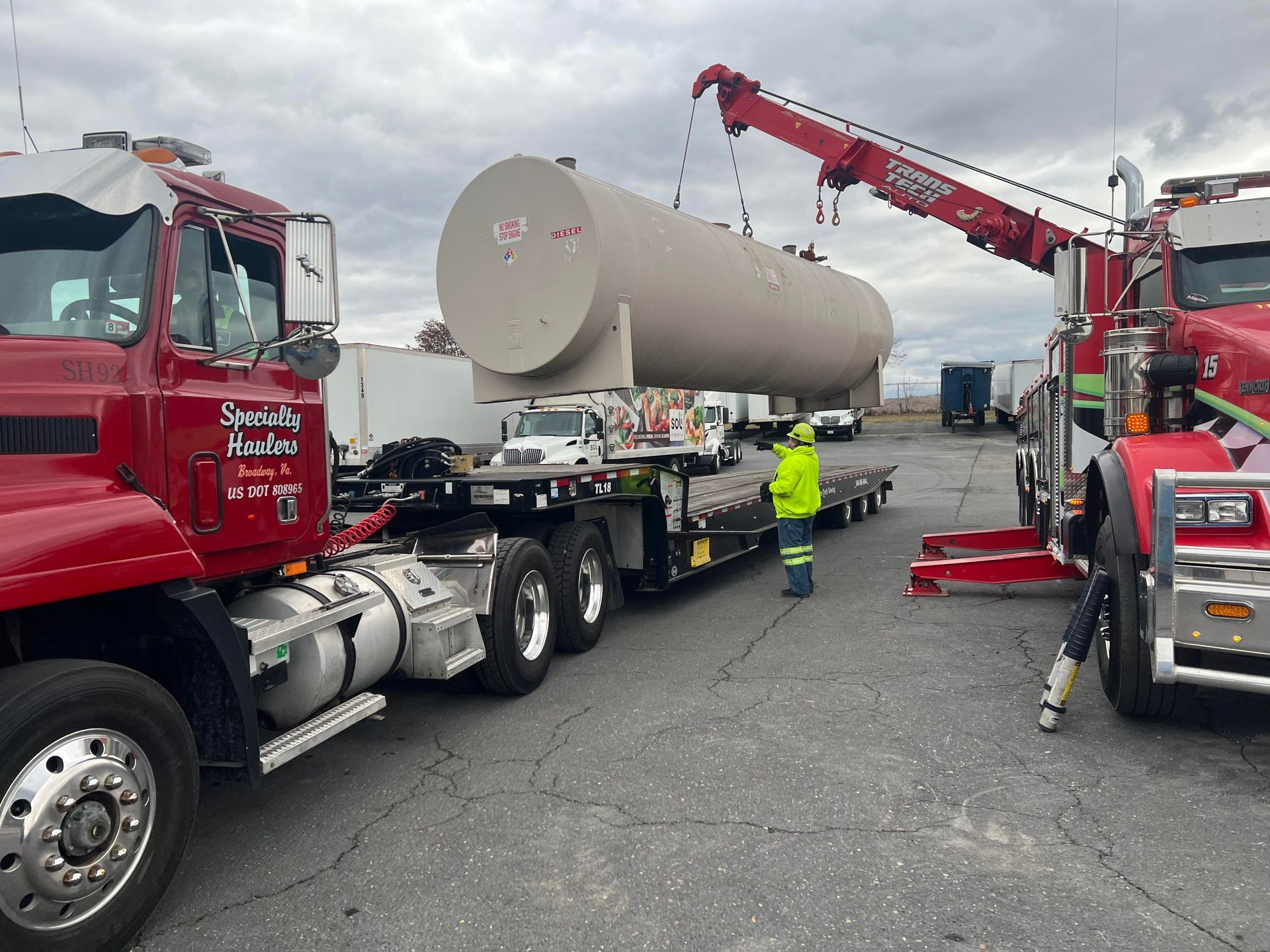 Red trucks and a crane load a large beige tank onto a flatbed in an industrial lot.