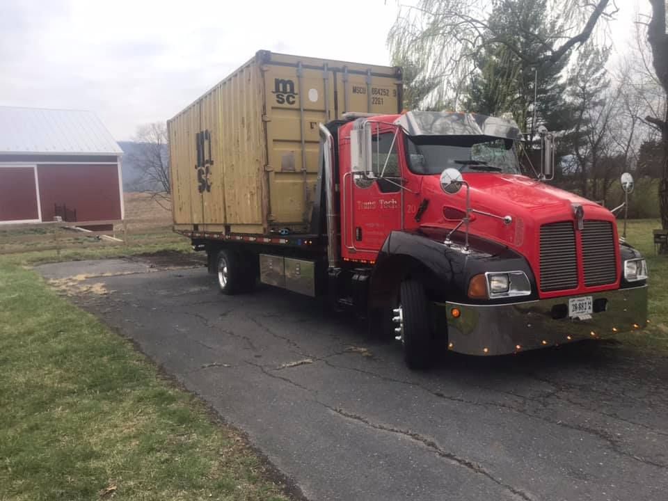 Red semi-truck hauling a tan shipping container on a driveway beside a red barn