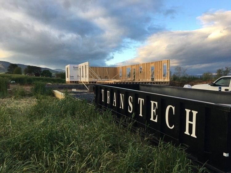 Sign reading Trans Tech in a grassy field with a wooden building under cloudy sky