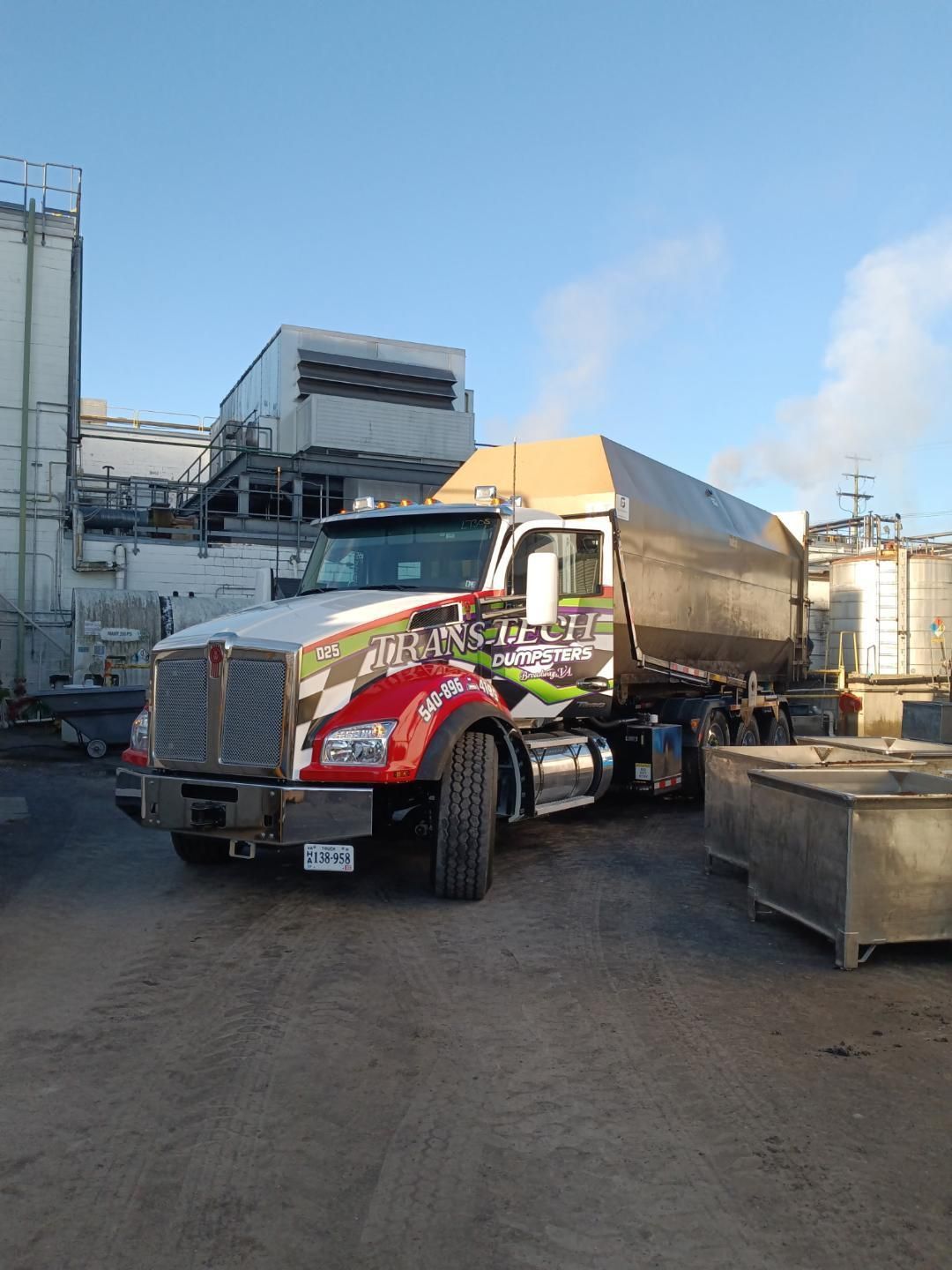 Red semi-truck parked in an industrial yard beside large tanks and machinery.
