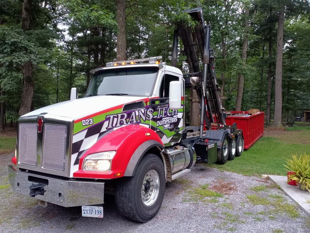White and red dump truck with a red trailer parked on gravel in a wooded area