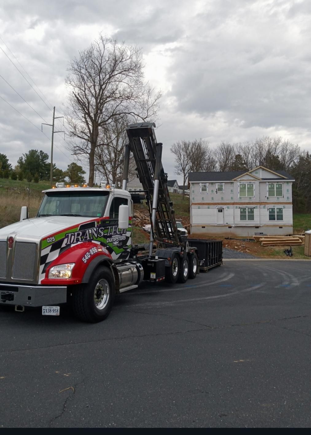 Tow truck hauling heavy equipment on a road beside a white building under cloudy skies
