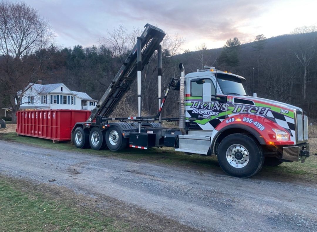 Tow truck hauling a red dumpster trailer on a gravel road at dusk