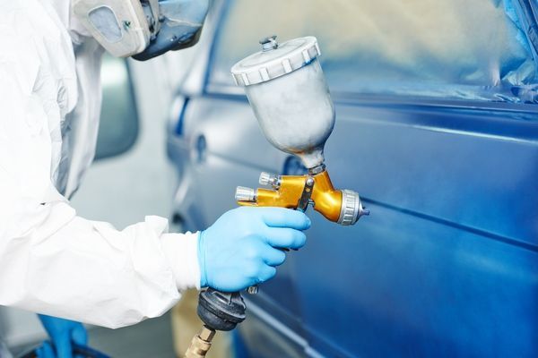Person in protective gear sprays blue paint on a car with a spray gun