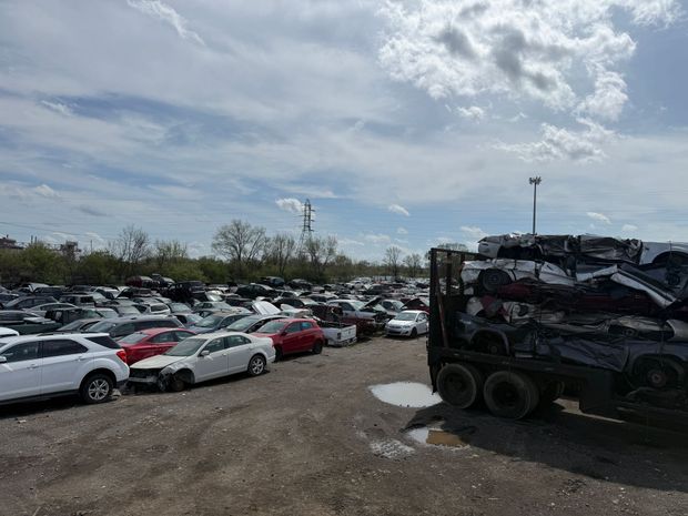 A large salvage yard filled with many parked, damaged cars, with a flatbed truck loaded with stacked vehicles on the right.