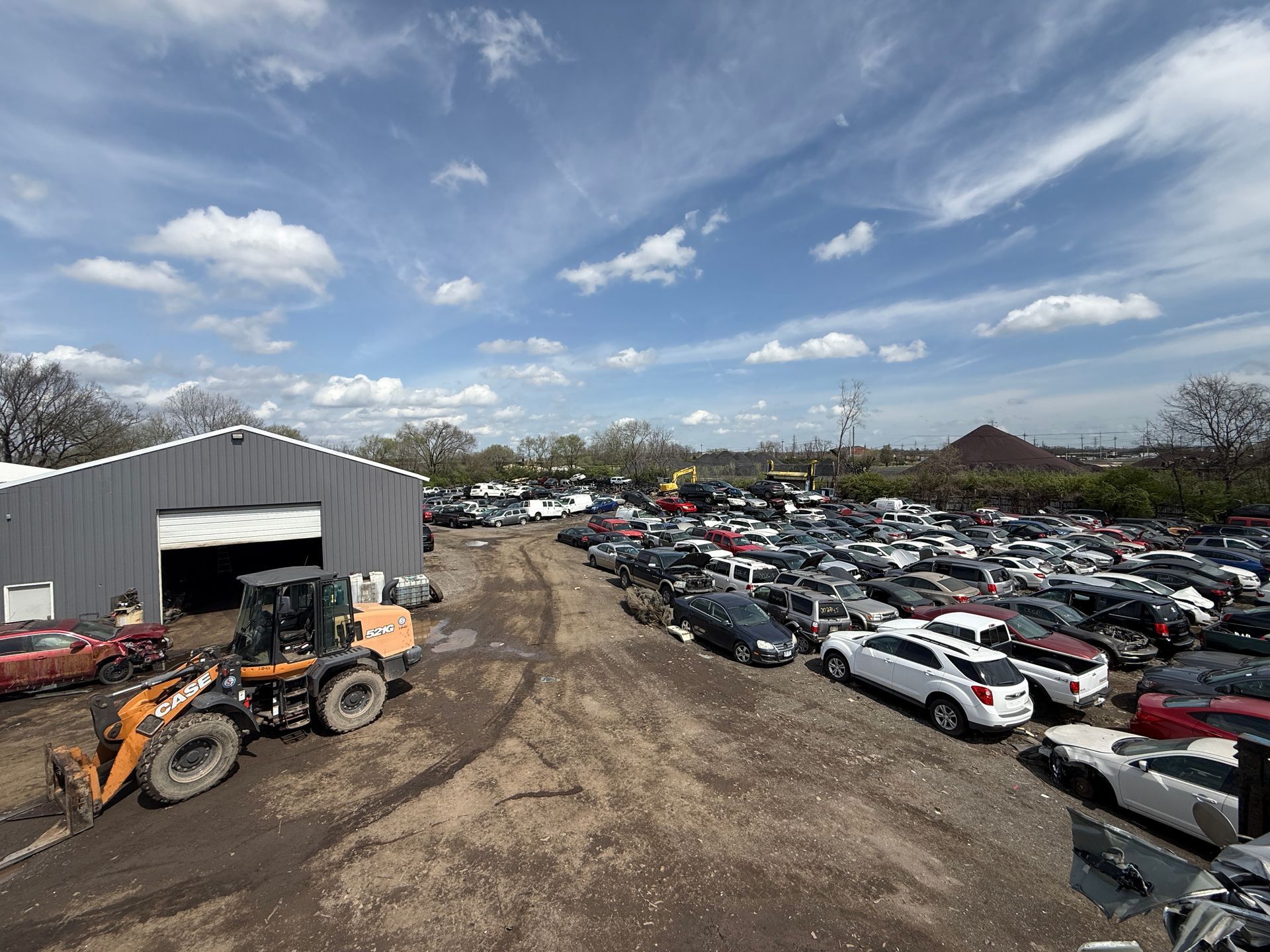 A construction vehicle sits on a muddy lot next to a gray warehouse, overlooking a vast salvage yard filled with cars.