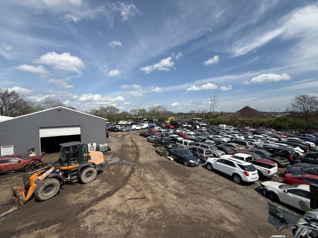 A large, muddy auto salvage yard featuring rows of parked vehicles and a front-end loader parked near a grey metal shed.