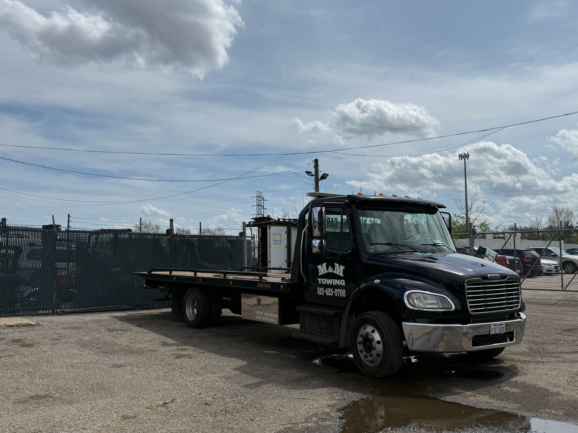 A black flatbed tow truck parked on a gravel lot under a partly cloudy sky.