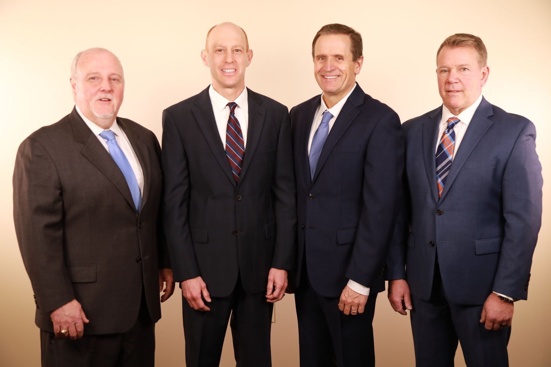 Four men in suits standing side-by-side, smiling slightly. Light background.