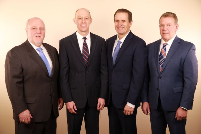 Four men in suits standing side-by-side, smiling slightly. Light background.