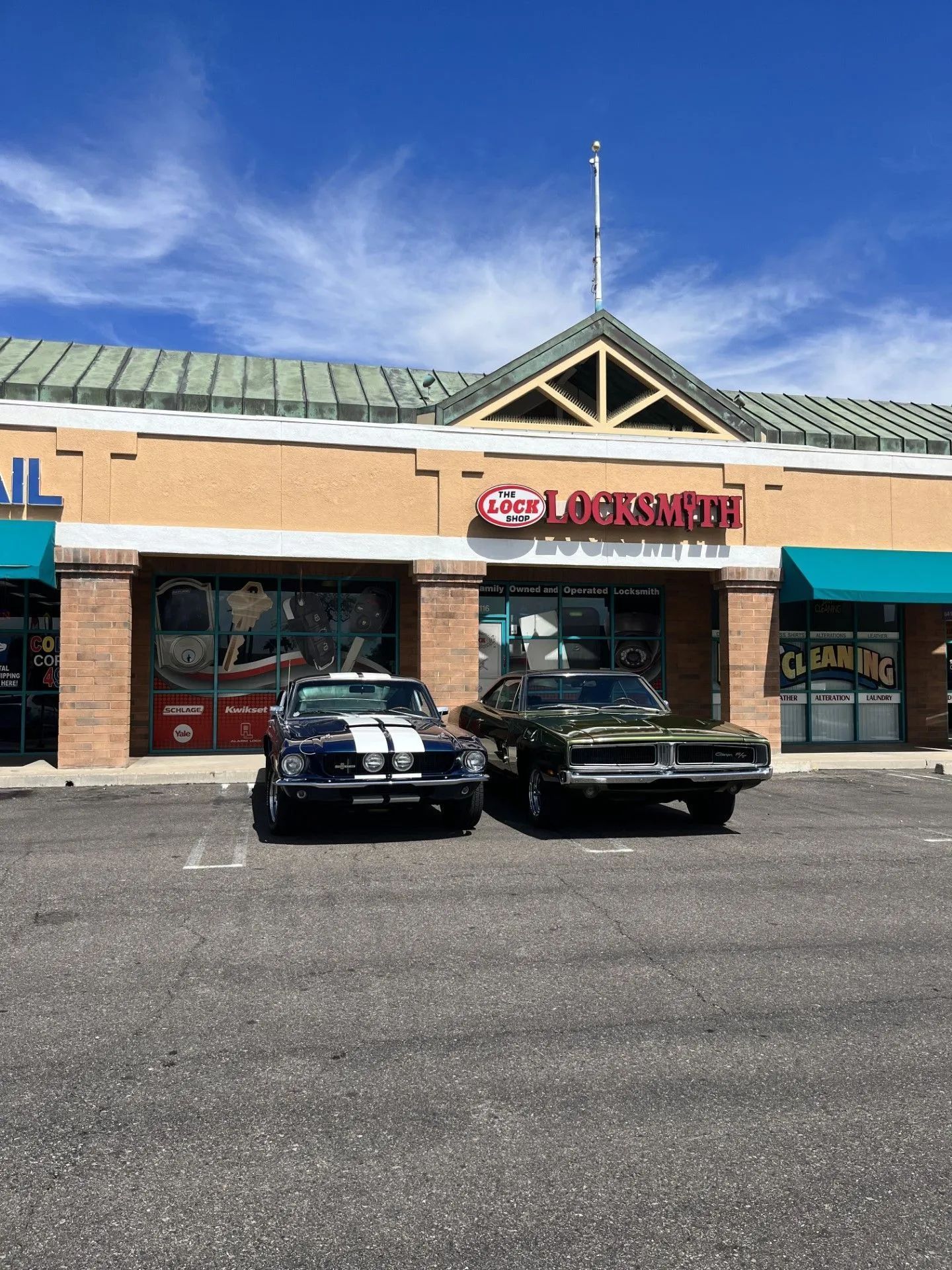 Two classic cars parked in front of a storefront: a blue Shelby Cobra and a green Dodge Charger.