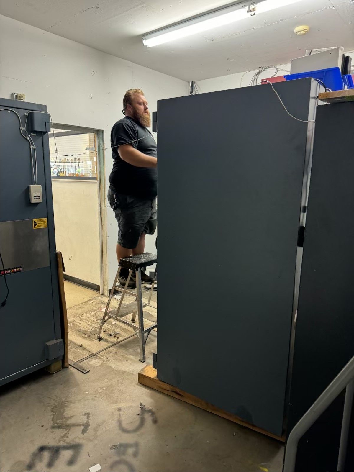 Man on a step stool in a cluttered room, near large gray cabinets.