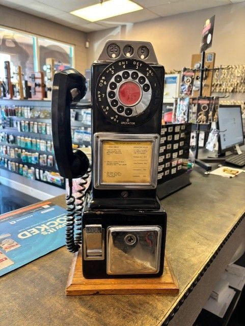 Black rotary payphone on a wooden base, inside a store with keys and other items.