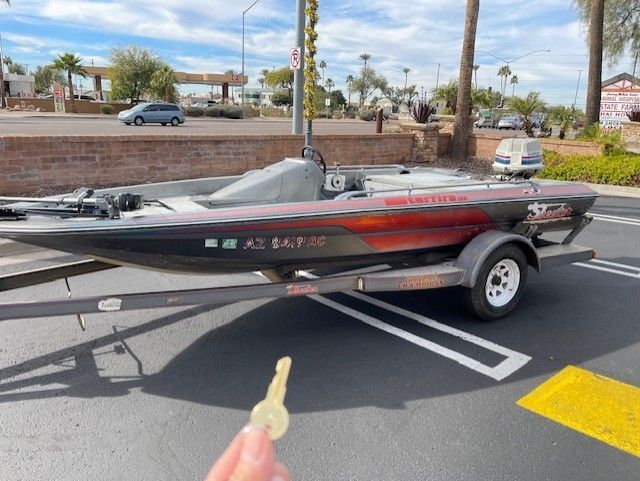 A hand holding a key in front of a bass boat on a trailer, parked on a street in Arizona.