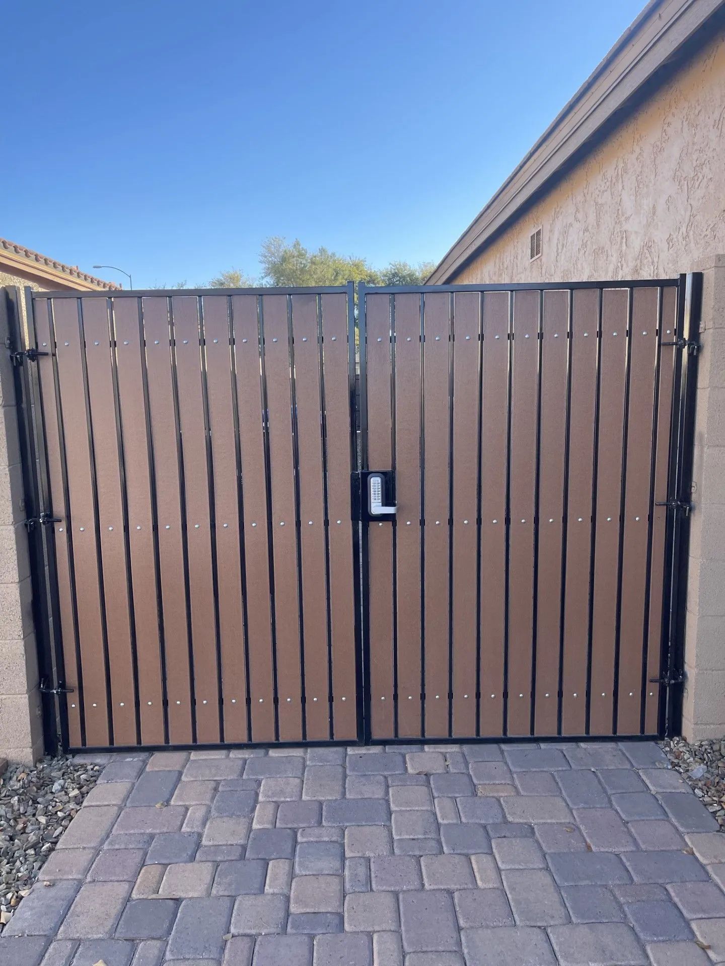 Two brown and black wooden gates on a brick path, against a tan wall, under a clear sky.