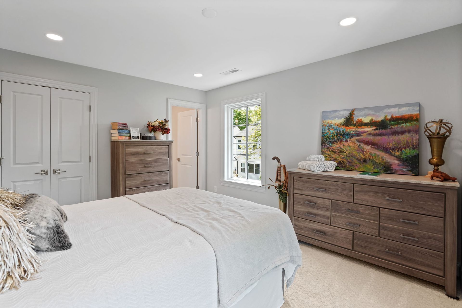 Bedroom with a white bed, wooden dressers, white doors, and a window with a painting above the dresser.
