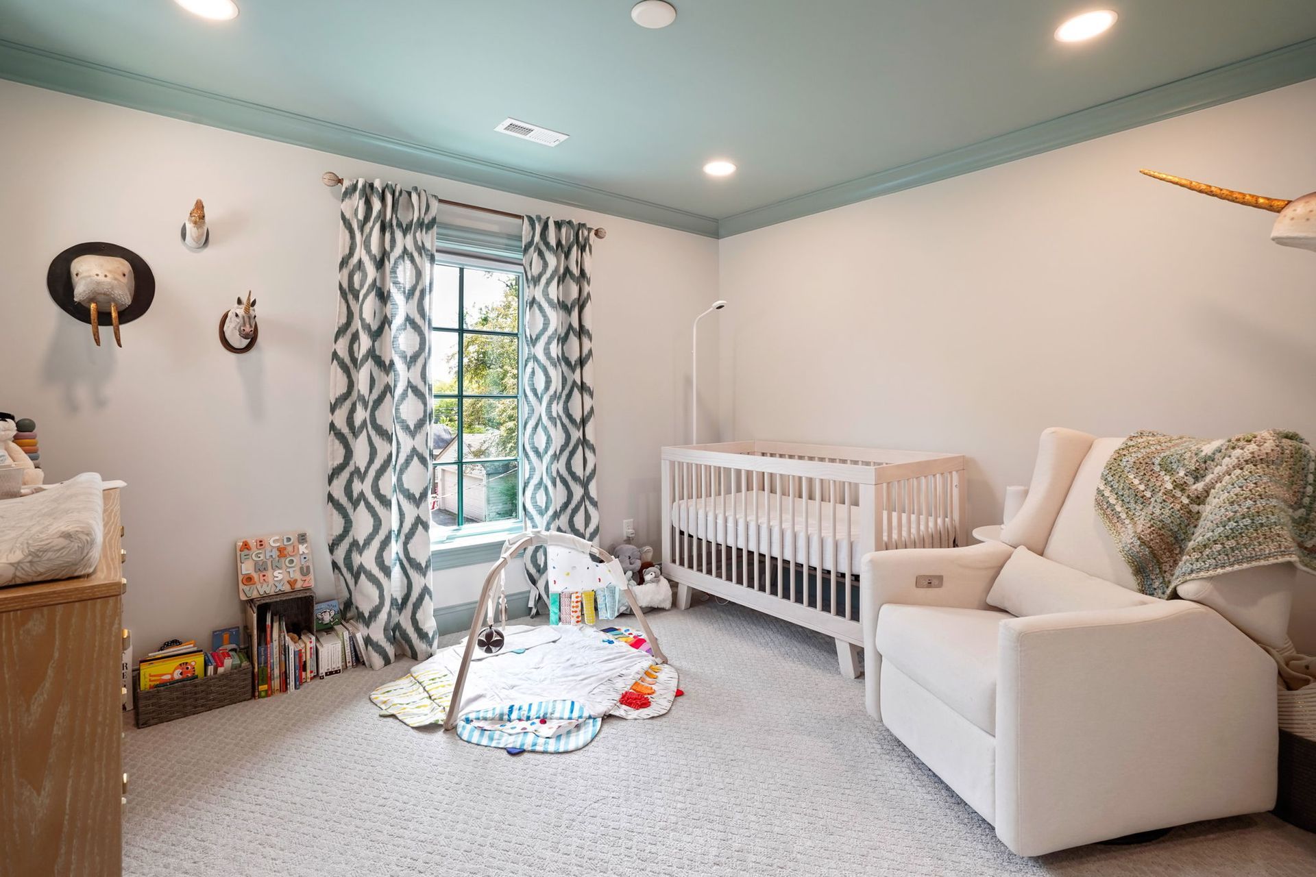 A nursery with a white crib, armchair, and play area. Teal ceiling and patterned curtains.