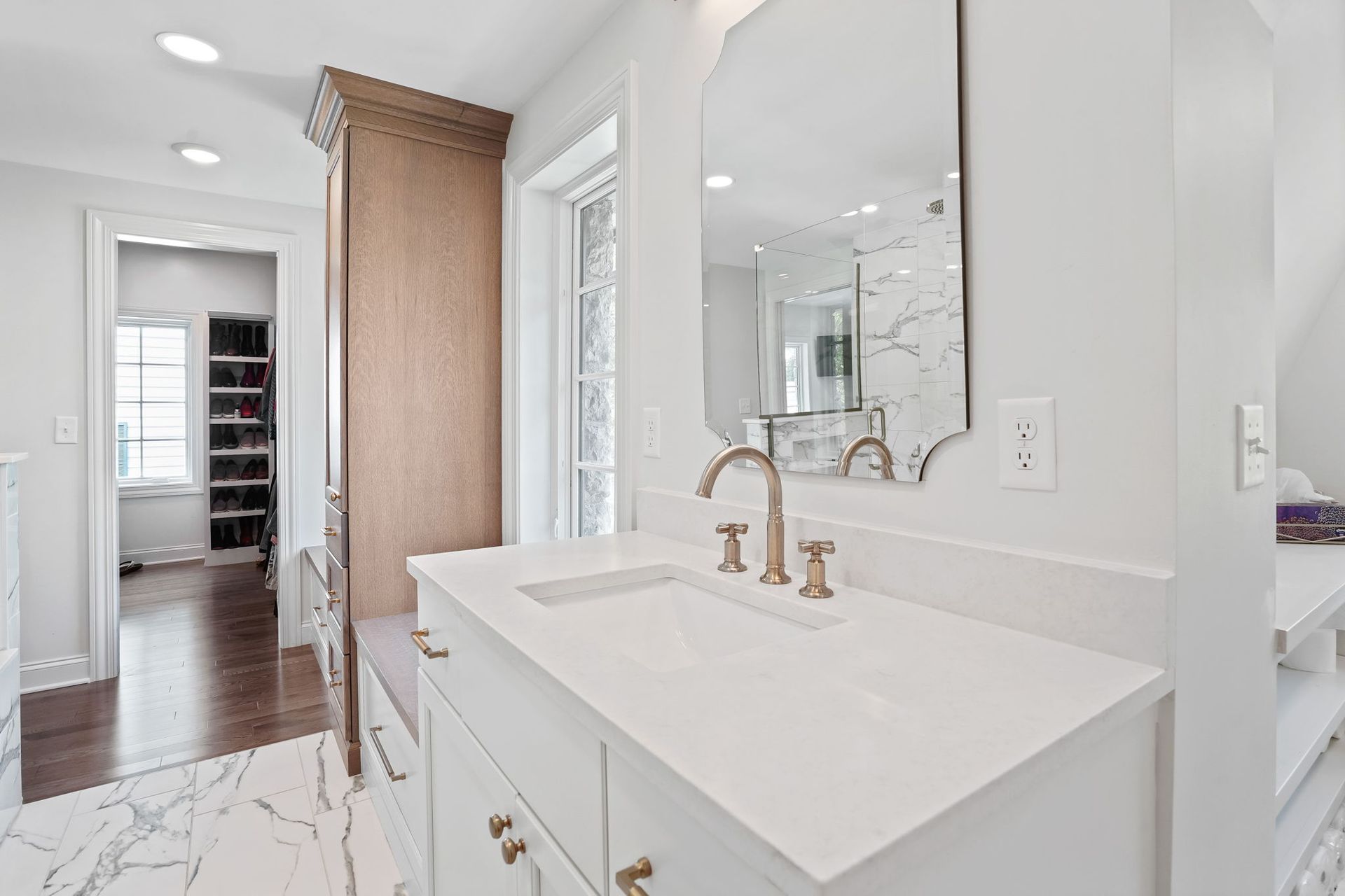 Bathroom with white countertop and sink, gold faucet, large mirror, and open closet in the background.