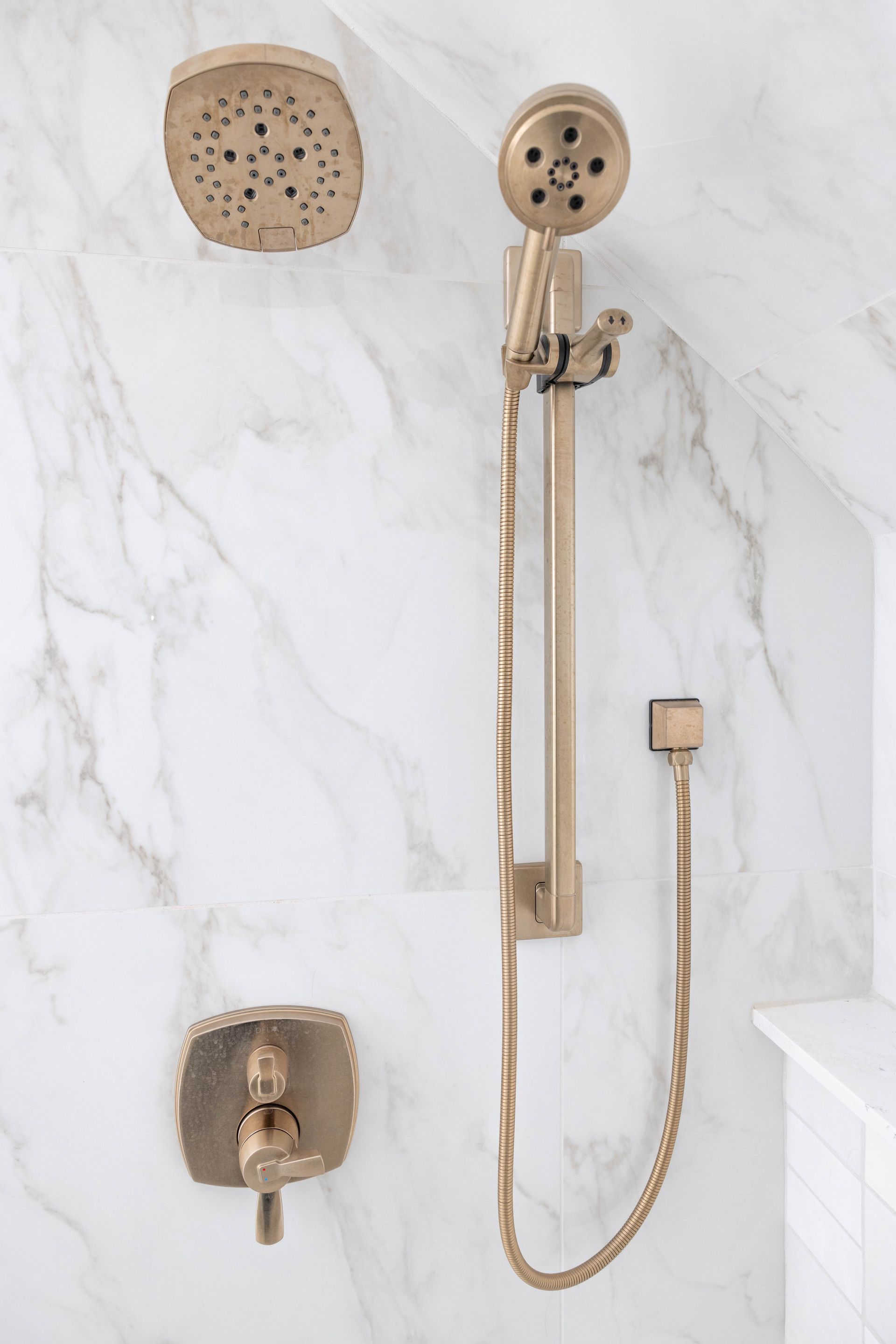 Gold shower fixtures mounted on white marble-look tile wall. Includes rain shower, hand shower, and water control.