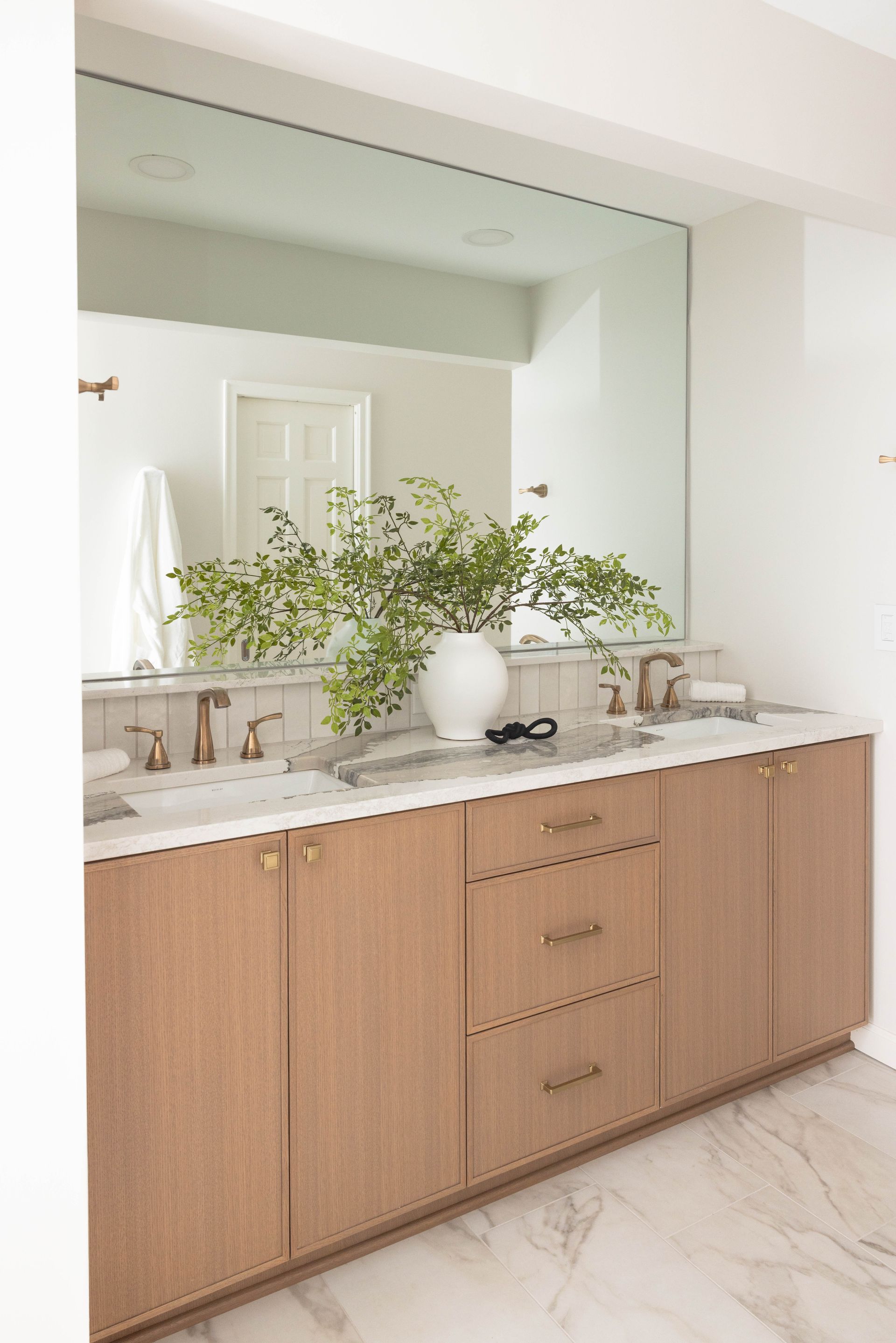 Bathroom vanity with light wood cabinets, marble countertop, large mirror, and vase of green branches.