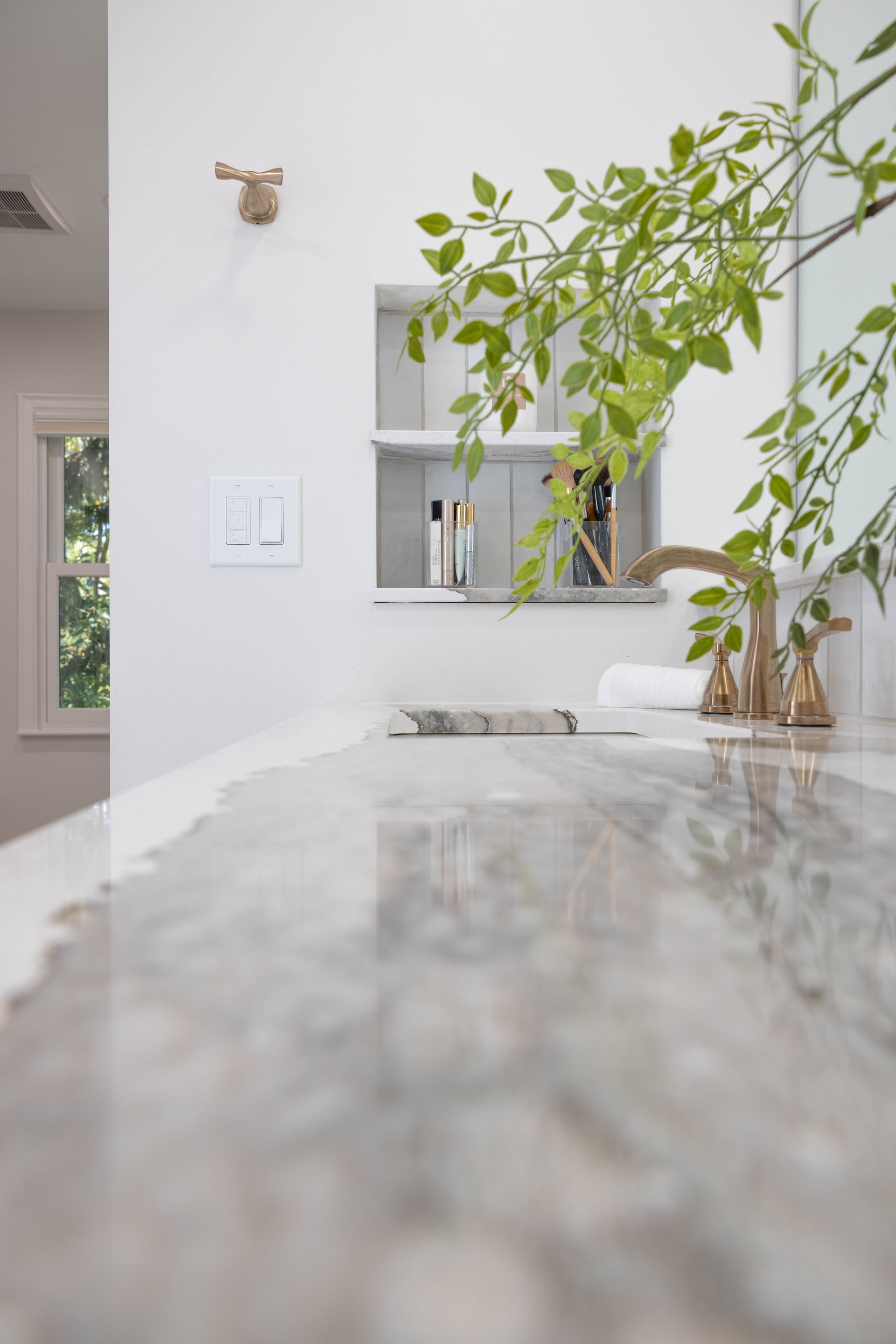 Bathroom countertop with gold faucet, built-in shelf, greenery, and window.